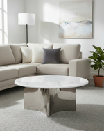 A modern living room scene featuring a round coffee table with a white marble top and a polished chrome base, positioned in front of a light beige sectional sofa with neutral throw pillows.
