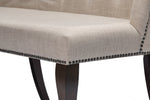 Close-up of an upholstered bench seat in a light beige textured fabric, with dark nailhead trim along the edge and dark wooden legs.