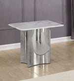 A modern side end table with a white marble top and a polished silver steel base, shown in a room setting with a gray paneled wall and light wood floor.