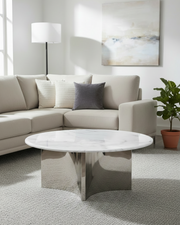A modern living room scene featuring a round coffee table with a white marble top and a polished chrome base, positioned in front of a light beige sectional sofa with neutral throw pillows.