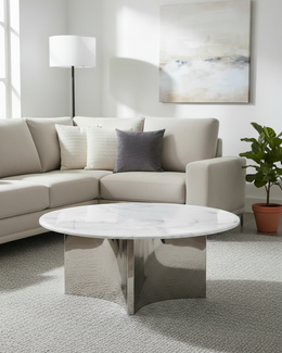 A modern living room scene featuring a round coffee table with a white marble top and a polished chrome base, positioned in front of a light beige sectional sofa with neutral throw pillows.