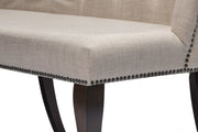 Close-up of an upholstered bench seat in a light beige textured fabric, with dark nailhead trim along the edge and dark wooden legs.